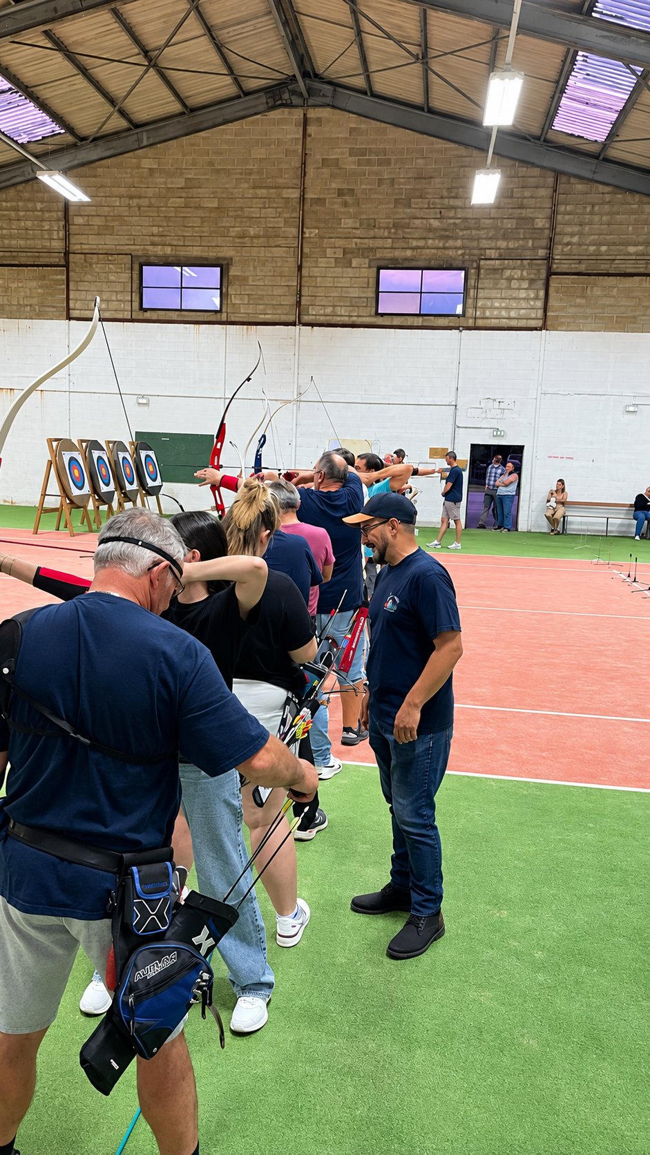 Un groupe d'archers en tenue sombre tire à l'arc vers des cibles dans une grande salle couverte, sous le regard d'un homme coiffé d'une casquette.
