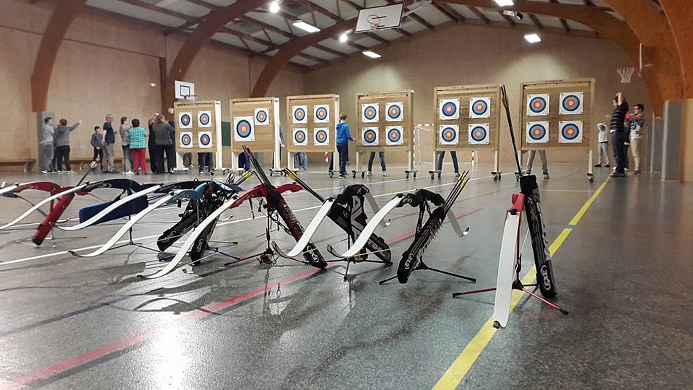 Dans un gymnase aux charpentes en bois, plusieurs arcs posés sur leurs supports font face à des cibles de tir à l'arc, devant des archers rassemblés.