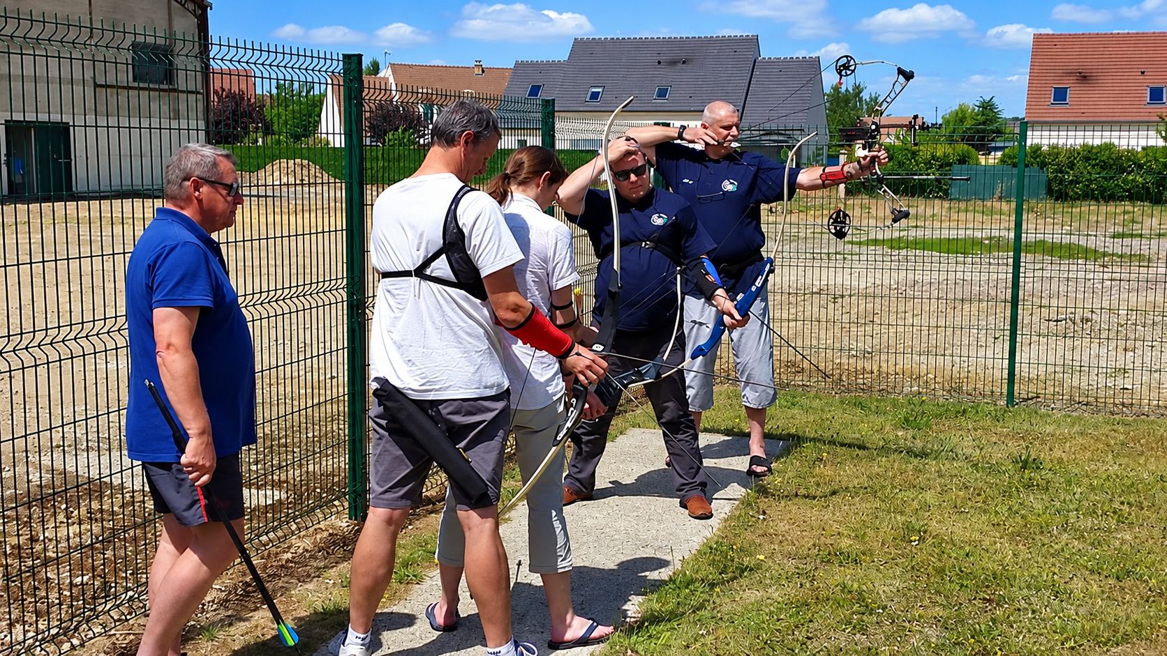 Quatre personnes pratiquent le tir à l'arc sur un terrain en plein air, devant une clôture verte, avec des arcs classiques et à poulies.