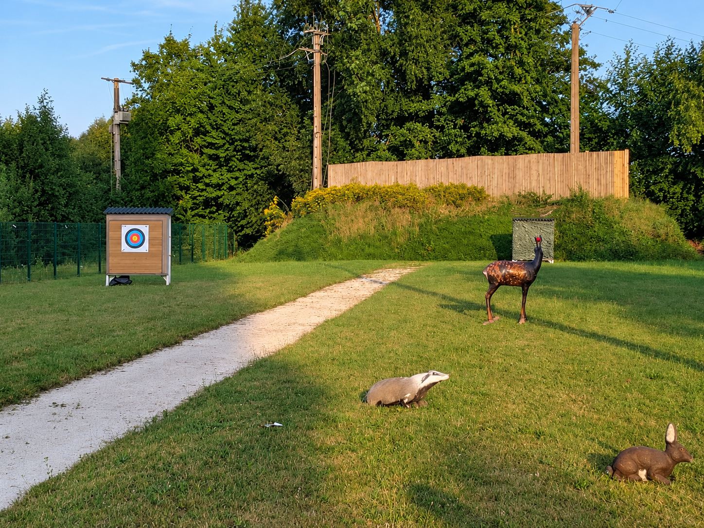 Sur un terrain herbeux au coucher du soleil, un chemin de gravier longe des cibles 3D en forme de blaireau, de cerf et de lièvre, avec une cible ronde sur un abri en bois au fond.