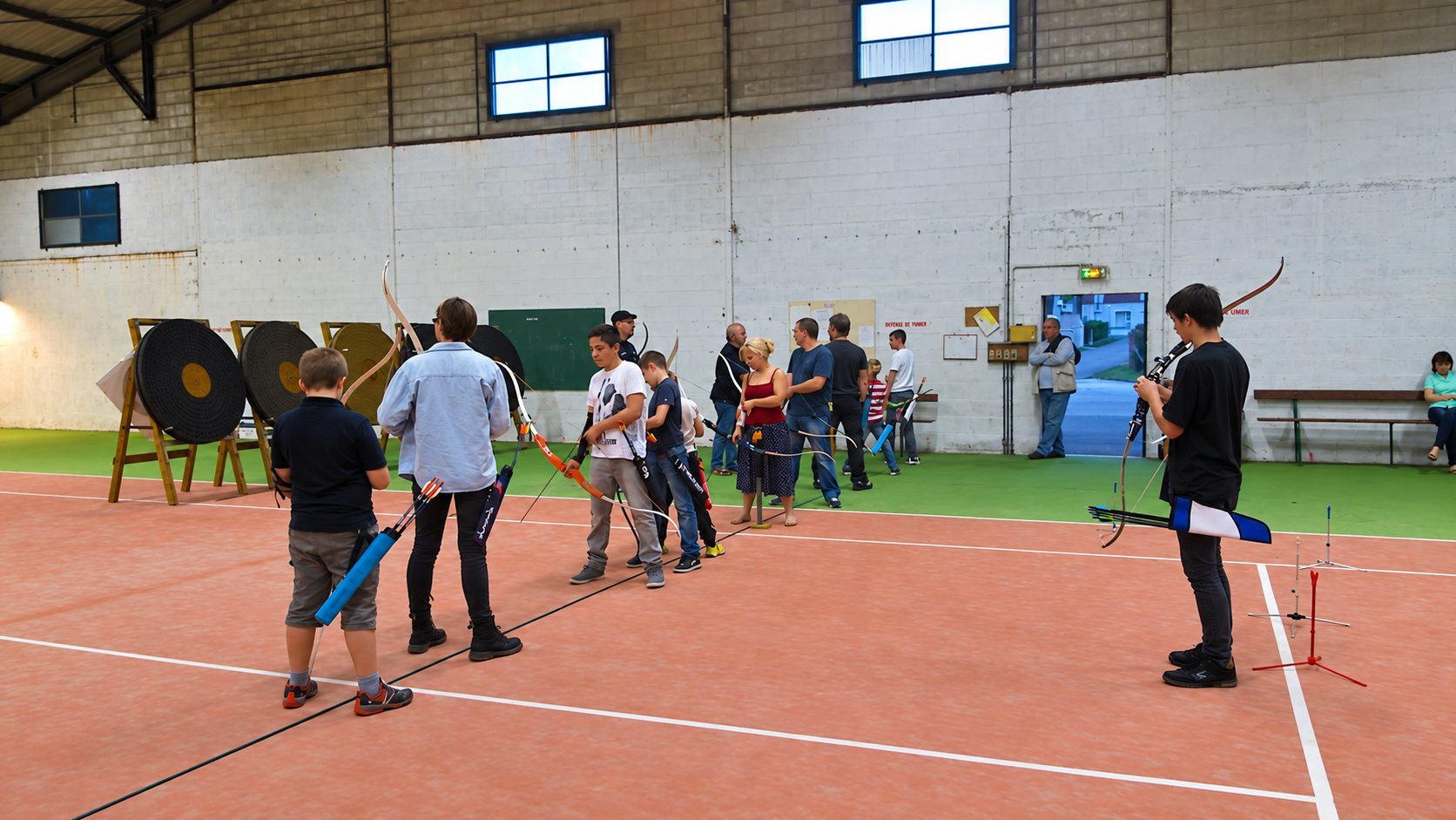 Des enfants et des adultes alignés avec leurs arcs devant des cibles lors d'une séance de tir à l'arc dans une salle couverte.